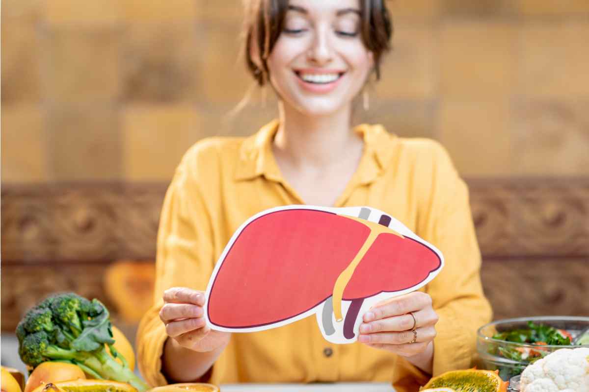 ragazza sorridente con una sagoma a forma di fegato in mano e alimenti che lo depurano sul tavolo
